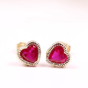 Pode incluir: Um par de brincos de coração de ouro com detalhes de pedras preciosas vermelhas.