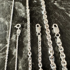 May include: Five silver chains of varying thicknesses and link styles. The chains are laid out horizontally on a dark gray marble surface.