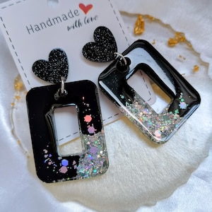 Peut inclure: Boucles d'oreilles pendantes composées de clous en forme de cœur noir attachés à des cadres rectangulaires noirs brillants. La partie inférieure de chaque rectangle est remplie de paillettes argentées irisées et de sequins hexagonaux. Les boucles sont présentées sur une carte blanche avec le texte Handmade with love.