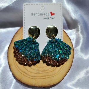 May include: Handmade seashell earrings with teal and bronze glitter, attached to gold-coloured studs. The earrings are displayed on a wooden disc. The tag reads "Handmade with love."