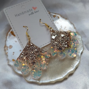 May include: A pair of fan-shaped dangle earrings featuring a gold glitter top section that transitions into a translucent white base filled with iridescent blue and silver glitter. The earrings hang from gold-toned hooks on a white display card.