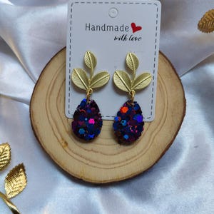 Peut inclure: Boucles d'oreilles faites à la main avec des détails de feuilles dorées et des pendentifs en forme de goutte remplis de paillettes colorées. Elles sont présentées sur une tranche de bois avec l'inscription "Handmade with love".
