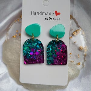 May include: A pair of handmade earrings with a teal top and a glittery teal and purple arch design. The earrings are attached to a white card with the text "Handmade with love".
