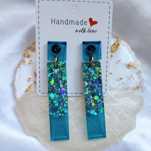 Può includere: Un paio di orecchini pendenti con un perno quadrato in resina color ottanio collegato da un anellino dorato a un lungo pendente rettangolare in resina. La parte inferiore è riempita di glitter blu e verdi. Il testo Handmade with love è stampato sul cartoncino bianco.