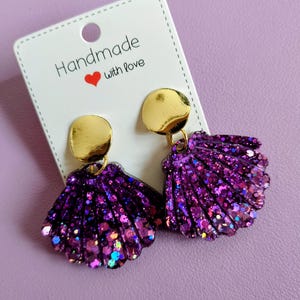 Peut inclure: Une paire de boucles d'oreilles coquillage faites à la main, avec des paillettes violettes et des clous circulaires dorés. Les boucles d'oreilles sont présentées sur une carte blanche avec l'inscription "Handmade with Love" et un cœur rouge.