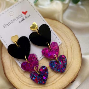May include: A pair of dangle earrings with three heart-shaped charms. The top charm is black, the middle charm is pink with iridescent glitter, and the bottom charm is purple with iridescent glitter. The charms are connected with gold-colored hoops.