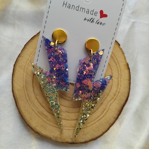 Puede incluir: Pendientes de rayo hechos a mano con purpurina. Los pendientes presentan una mezcla de purpurina morada, rosa y dorada en resina. Se exhiben sobre una rodaja de madera con la etiqueta "Handmade with love."