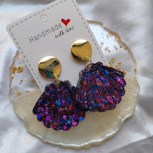 Op de afbeelding: Handgemaakte schelpvormige oorbellen met goudkleurige schijfjes. De oorbellen zijn donkerpaars met blauwe, roze en oranje glitter. De kaart zegt "Handmade with love."