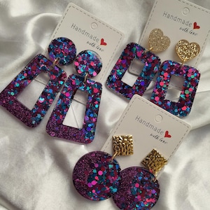 Puede incluir: Una colección de pendientes hechos a mano con varios diseños. Algunos pendientes presentan purpurina en tonos morados, azules y rosas. Otros tienen formas de corazón y cuadrado dorados. Cada par se muestra en una tarjeta que dice "Handmade with love."