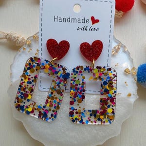Może przedstawiać: Ręcznie robione kolczyki z czerwonymi brokatowymi sercami i przezroczystymi prostokątami wypełnionymi kolorowym konfetti. Na karcie widnieje napis "Handmade with love".