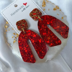Peut inclure: Une paire de boucles d'oreilles pendantes avec un clou carré à paillettes dorées et un grand pendentif en forme de U avec des paillettes rouge vif. Les boucles sont fixées sur un support blanc portant l'inscription Handmade with love.