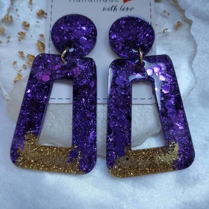 Könnte beinhalten: Hängeohrringe mit einem kreisförmigen Stecker und einem rechteckigen Anhänger. Die Stücke bestehen aus violettem Harz, gefüllt mit violettem Glitzer und einem goldenen Glitzerakzent am unteren Rand. Die Ohrringe sind auf einer Karte mit Handmade with love befestigt.
