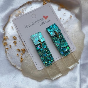 Può includere: Un paio di orecchini pendenti con un elemento superiore quadrato e uno inferiore rettangolare collegati da un anello in metallo dorato. Gli elementi sono in resina trasparente con glitter color verde acqua, blu e verde. Gli orecchini sono fissati su un cartoncino bianco con la scritta Handmade with love.