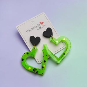 Peut inclure: Une paire de boucles d'oreilles sur un support blanc avec le texte Handmade with love. Chaque boucle présente un clou en forme de cœur noir uni attaché à un pendentif plus grand en forme de cœur évidé, vert lime avec des paillettes noires et dorées.