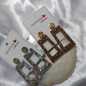 May include: Two pairs of rectangular dangle earrings. One pair is gold glitter, the other is iridescent glitter. Each pair is attached to a white card that reads "Handmade with love" with a red heart.