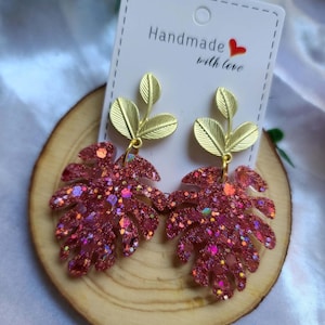 Puede incluir: Pendientes hechos a mano con diseño de hoja. Los pendientes tienen un tallo de hoja dorado y un colgante de hoja rosa brillante. Se exhiben en una rodaja de madera con una etiqueta que dice "Handmade with love."