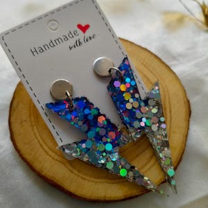 May include: A pair of handmade lightning bolt earrings, adorned with iridescent glitter in blue, silver, and other colours. The earrings are attached to silver posts and presented on a white card with the text "Handmade with love."