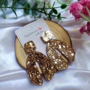 Può includere: Orecchini dorati con glitter, con parte superiore quadrata e inferiore ad arco. Gli orecchini sono su una fetta di legno con un'etichetta che dice "Handmade with love".