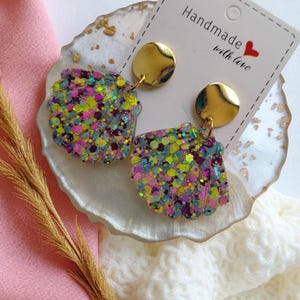 Peut inclure: Boucles d'oreilles faites à la main en forme de coquillage, avec des paillettes colorées et des clous dorés. Les boucles d'oreilles sont présentées sur une surface décorative blanche et dorée. Une étiquette indique "Handmade with love."