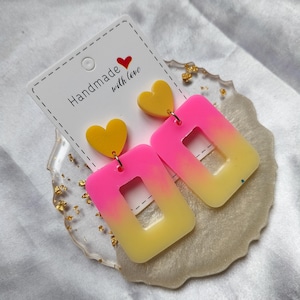 Puede incluir: Pendientes hechos a mano con diseño de corazón y rectángulo. Los pendientes presentan un corazón amarillo y un rectángulo degradado de rosa a amarillo. La tarjeta blanca dice "Handmade with love".