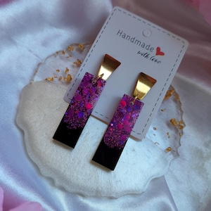 May include: A pair of rectangular earrings with a black base and a glittery purple top. The earrings have gold-colored metal accents and are displayed on a white card that reads "Handmade with love".