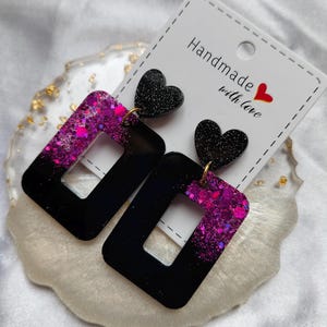 Puede incluir: Pendientes rectangulares negros y rosas con purpurina y un corazón en la parte superior. Los pendientes están sobre una tarjeta blanca que dice "Handmade with love". Los pendientes están sobre una superficie decorativa blanquecina.