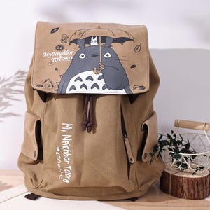 Puede incluir: Mochila de lona color beige con una ilustración de dibujos animados de Totoro sosteniendo un paraguas. La solapa tiene el texto "My Neighbor Totoro" y un cierre de cordón. La mochila tiene bolsillos laterales y una cremallera.