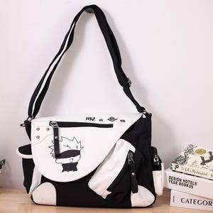 Puede incluir: Bolso mensajero en blanco y negro con correa para el hombro. El bolso presenta un gráfico de dibujos animados en la solapa delantera y múltiples bolsillos con cremallera. Las dimensiones del bolso son de aproximadamente 35 cm de ancho, 28 cm de alto y 10 cm de profundidad.