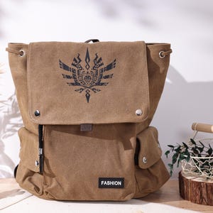 May include: A brown canvas backpack with a flap closure and two side pockets. The flap features a black graphic design and the word "FASHION" is printed on the bottom. The backpack has adjustable drawstring closures and silver metal accents.