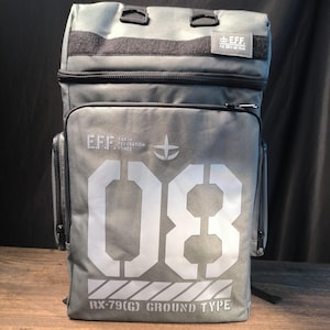 Könnte beinhalten: Grauer Rucksack mit dem Text "EFF EARTH FEDERATION FORCE" und der Zahl "08" in Weiß. Der Text "RX-79(G) GROUND TYPE" ist ebenfalls sichtbar. Der Rucksack hat eine obere Klappe und mehrere Taschen.