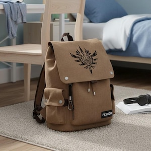 Może przedstawiać: Brązowy płócienny plecak z klapą i dwiema bocznymi kieszeniami. Klapa ma czarny wzór graficzny. Plecak ma brązowe paski i małą metkę z napisem "FASHION". Leży na jasnym dywanie.