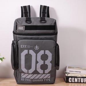 May include: A grey backpack with black straps and side pockets. The front displays the text "EFF" and the number "08" in a large, bold font. Below the number is the text "RX-79(G) GROUND TYPE". The backpack has a rectangular shape and a padded back.