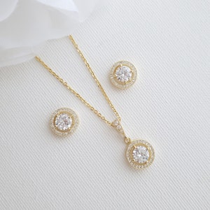 May include: A gold necklace and earrings set with a round pendant and matching earrings. Each piece features a large clear crystal surrounded by a halo of smaller crystals.