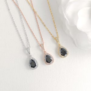 May include: Three pendant necklaces with teardrop-shaped black gemstones. The necklaces are silver, rose gold, and gold, each with a delicate chain and a halo of small, clear stones around the black gem. The pendants are displayed on a white surface.