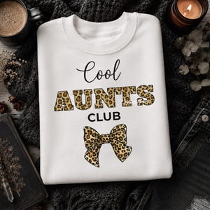 May include: A white crewneck sweatshirt featuring the black text Cool and CLUB, with the word AUNTS in a brown and black leopard print pattern. Below the text is a graphic of a bow with the same leopard print design on a white background.