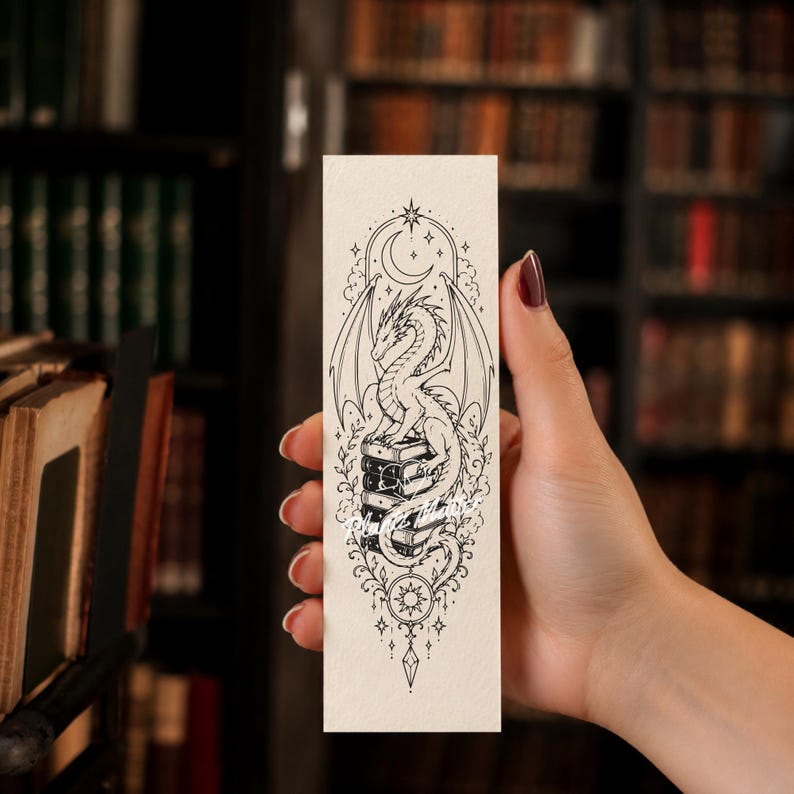 May include: A rectangular bookmark featuring a black and white line art illustration of a dragon perched atop a stack of books. The design includes a crescent moon, stars, and a sun motif with decorative filigree elements on a cream background.