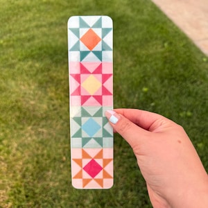 May include: A rectangular bookmark with a colorful quilt block design. The design features alternating star patterns in shades of orange, teal, pink, yellow, and blue. The bookmark is held against a background of green grass.