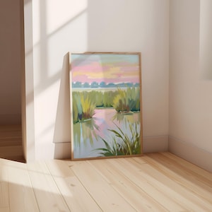 Op de afbeelding: Een ingelijst landschapsschilderij toont een moeraslandschap met een pastelkleurige zonsondergang. Het kunstwerk toont groene riet, een reflecterend water en een lucht met roze, gele en blauwe tinten. De houten lijst complementeert het zachte kleurenpalet van het kunstwerk.
