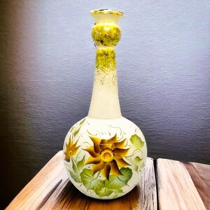 Può includere: Un vaso in ceramica bianca con corpo rotondo e collo lungo e sottile. Il vaso è decorato con girasoli gialli dipinti a mano e foglie verdi.