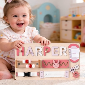 Könnte beinhalten: Holz-Aktivitätstafel mit dem Namen "HARPER" in rosa und weißen Buchstaben. Die Tafel verfügt über interaktive Elemente wie Perlen, einen Reißverschluss, eine Schnalle und Zahnräder. Ein lächelndes Baby hält die Tafel.