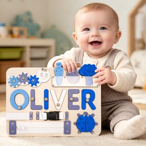 May include: Wooden activity board with the name "OLIVER" in blue and purple letters. Features include gears, vegetable shapes, a zipper, a buckle, and a ladybug. The board is designed for early childhood development.