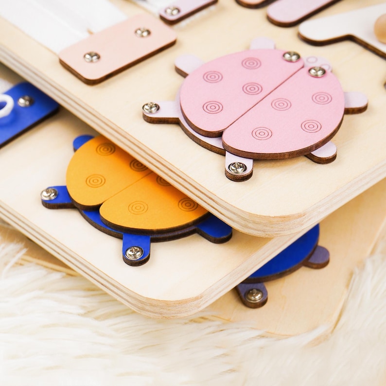May include: Wooden educational toy with colorful, geometric shapes and ladybug designs. The toy features a light wood base with attached pieces in shades of pink, orange, and blue. The ladybug pieces are secured with silver screws.
