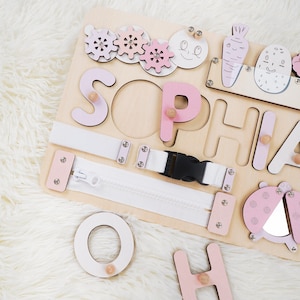 May include: Wooden activity board with the name "SOPHIA" spelled out in pink letters. Features include a zipper, buckle, gears, and a ladybug design. The board is designed for educational play.