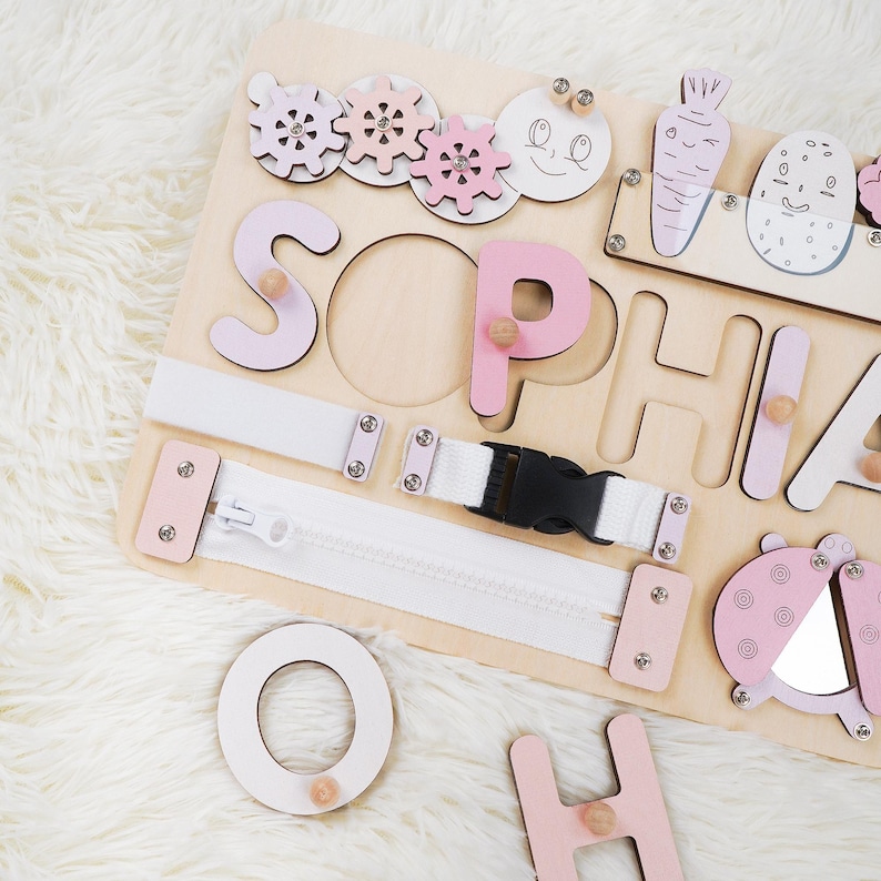 May include: Wooden activity board with the name "SOPHIA" spelled out in pink letters. Features include a zipper, buckle, gears, and a ladybug design. The board is designed for educational play.