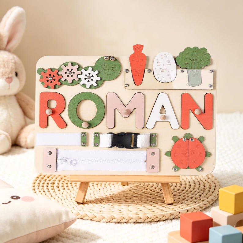 May include: Wooden educational toy with the name "ROMAN" spelled out in colorful letters. The board features various interactive elements like gears, a zipper, a buckle, and vegetable-shaped cutouts. It is designed for early learning and sensory exploration.