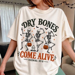 Peut inclure: Un t-shirt couleur crème avec un motif de trois squelettes dansants portant des seaux citrouilles orange et une pierre tombale. Le texte indique DRY BONES, COME ALIVE et EZEKIEL 37:5 en noir et orange, avec des petites chauves-souris et étoiles.
