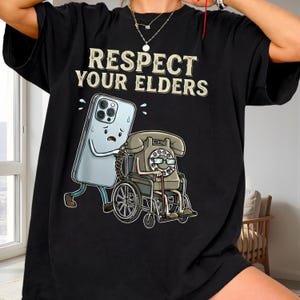May include: Black t-shirt with the text "RESPECT YOUR ELDERS" in a vintage font. The graphic shows a cartoon smartphone pushing a vintage telephone in a wheelchair. The smartphone has a worried expression.