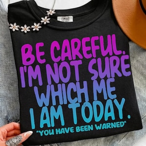 Könnte beinhalten: Schwarzes Rundhals-T-Shirt mit dem Text "BE CAREFUL. I'M NOT SURE WHICH ME I AM TODAY. *YOU HAVE BEEN WARNED*". Der Text ist in einem Farbverlauf aus Lila, Blau und Türkis gehalten. Eine Blumenkette und ein brauner Hut sind ebenfalls zu sehen.