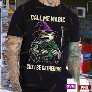 Puede incluir: Camiseta negra con un diseño de una rana con un sombrero de mago morado y una túnica, sosteniendo dinero. El texto "CALL ME MAGIC CUZ I BE GATHERING" está impreso. Un diseño humorístico sobre magia y riqueza.