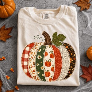 May include: A cream-coloured short-sleeve t-shirt featuring a large, central pumpkin graphic composed of various fabric-style patches in red, green, orange, and brown with floral, plaid, and herringbone patterns. The shirt has a Comfort Colors label.